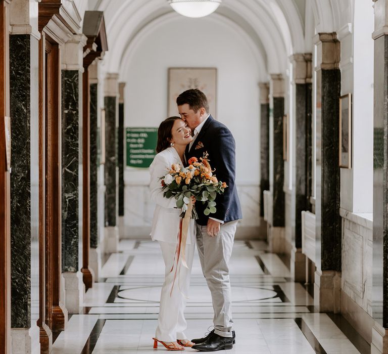 Bride and groom portrait in Islington Town Hall with bride in a jumpsuit and orange shoes holding an orange flower bouquet 