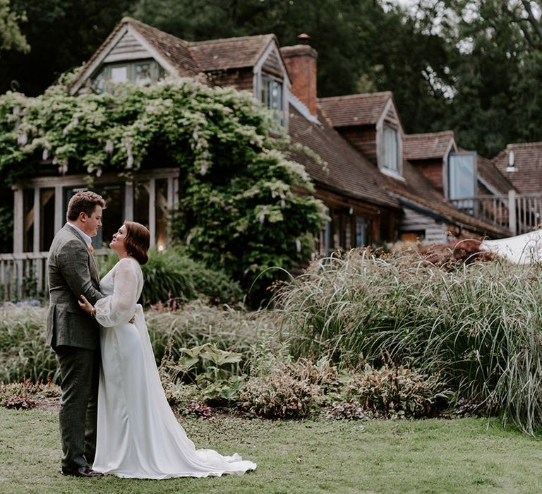 Bride in separates embracing her groom in a grey check suit in front of their wedding venue 