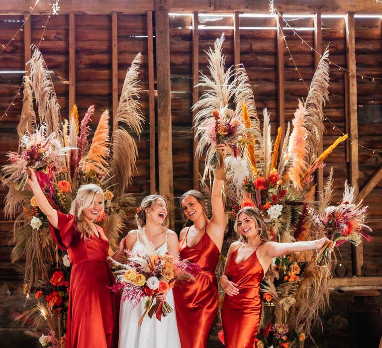 Laughing bride in lace wedding dress with train and veil stands in front of rose and pampas grass rustic floral installation with bridesmaids in orange satin bridesmaid dresses