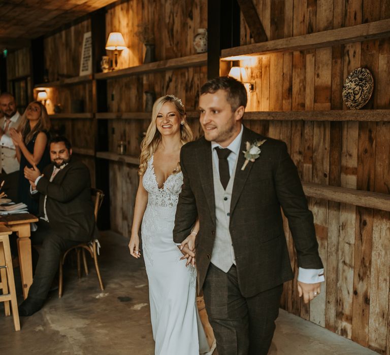 Groom in three piece brown suit with white rose buttonhole walks through barn wedding reception holding hands with bride in lace wedding dress