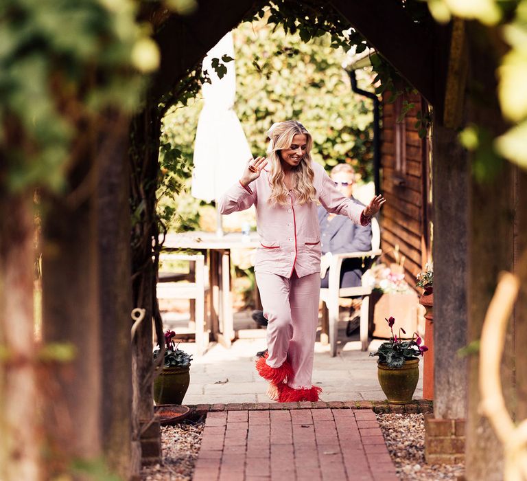 Bride with curled blonde hair wearing pink pyjamas with red fur trim dances in garden 