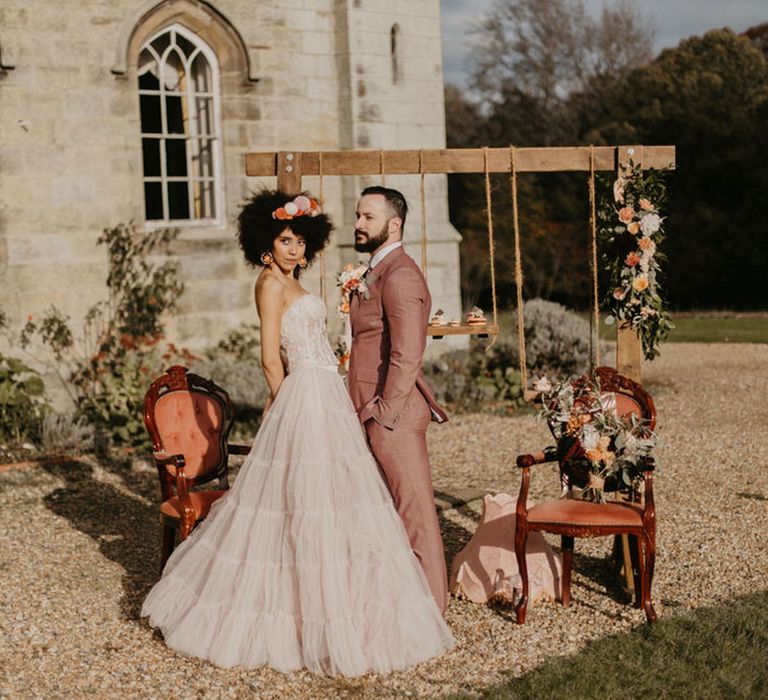 Wooden frame with hanging cake stands, ornate chairs and Black bride with afro hair in pink bridal separates with her groom in a pink suit 