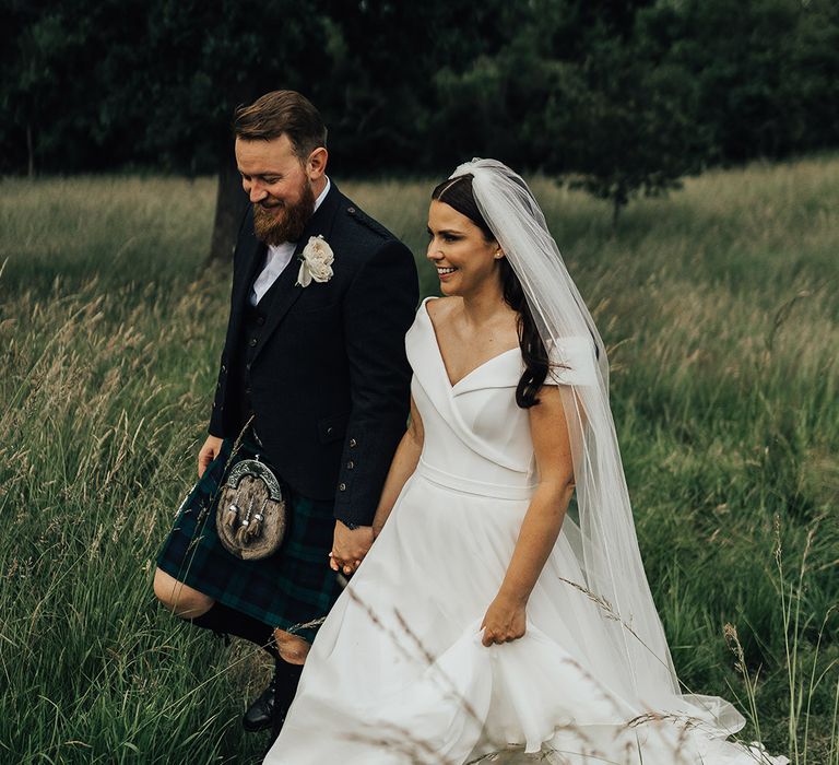 Bride in a Suzanne Neville Bardo wedding dress and long veil walking through the fields with her groom in a tartan kilt 