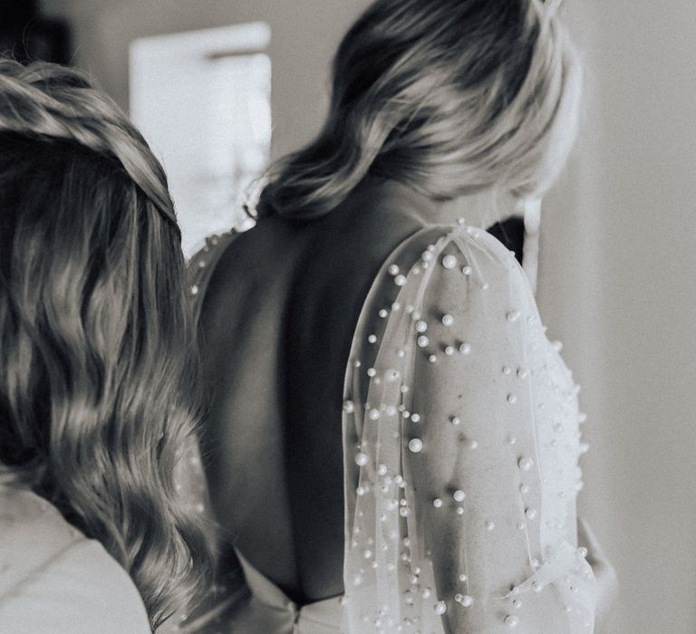 Bride in pearl wedding dress with sleeves has her dress buttoned up by bridesmaid before wedding