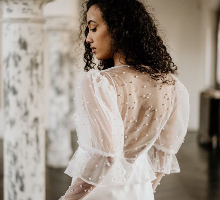 Satin low back wedding dress complete with sheer pearl jacket with layered sleeves worn by bride with curly hair 