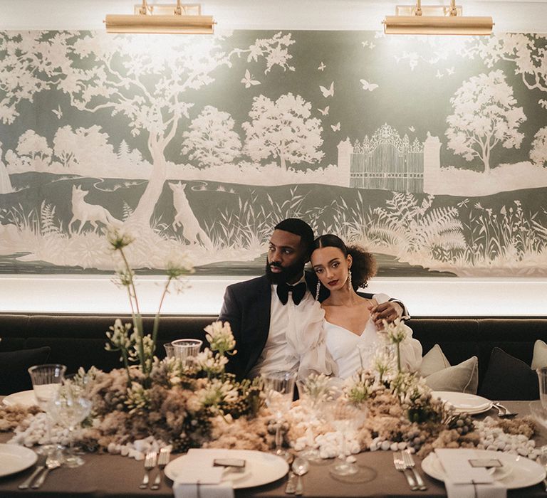 Black bride and groom sitting at their stylish wedding reception at Mayfair Townhouse with green and white flowers 