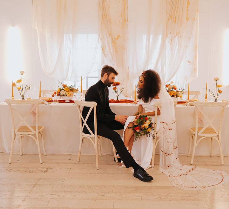 Black bride in an off the shoulder wedding dress with patterned veil sitting at her reception table with her groom in a all-black suit and top 