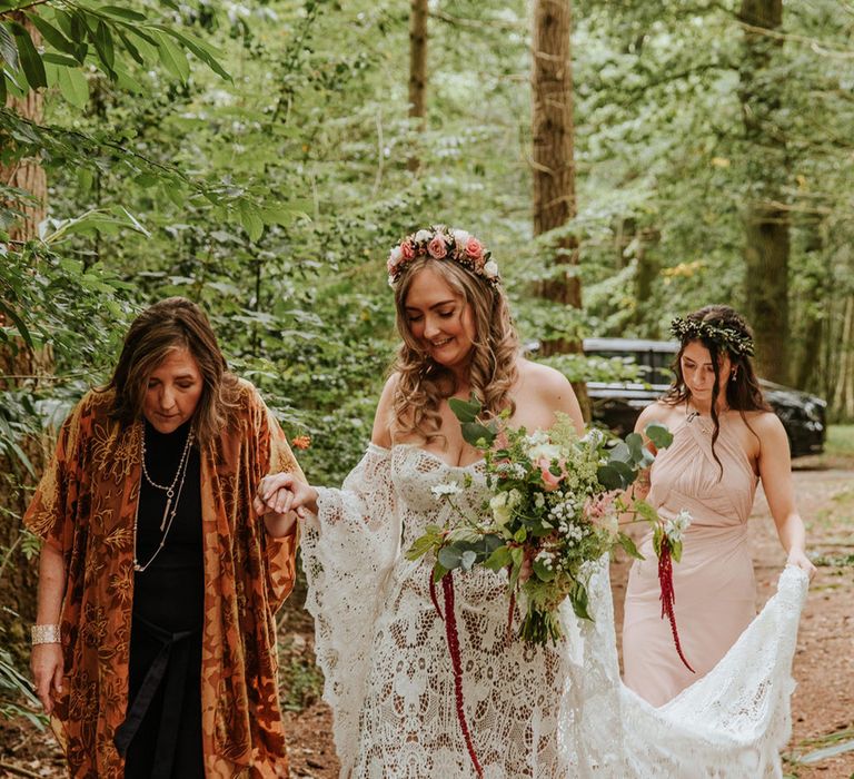 Bride in medieval style lace wedding dress and flower crown walks with mother as bridesmaid in pink dress holds up the train