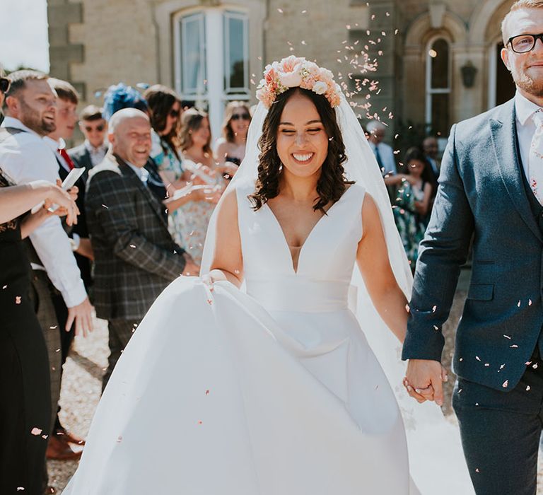 Groom in three piece blue suit with white floral tie with bride wearing flower corn smile as they have confetti exit 