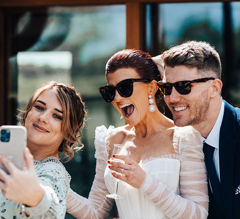 Bride takes a selfie with wedding guests wearing sunglasses and statement silver earrings 