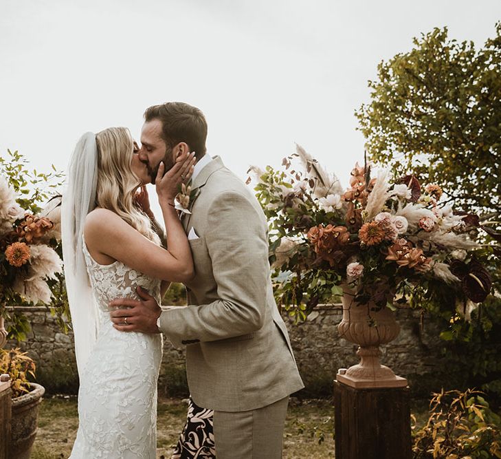 Bride & groom kiss between extravagant floral bouquets complete with tones of burnt orange, whites and pampas grass