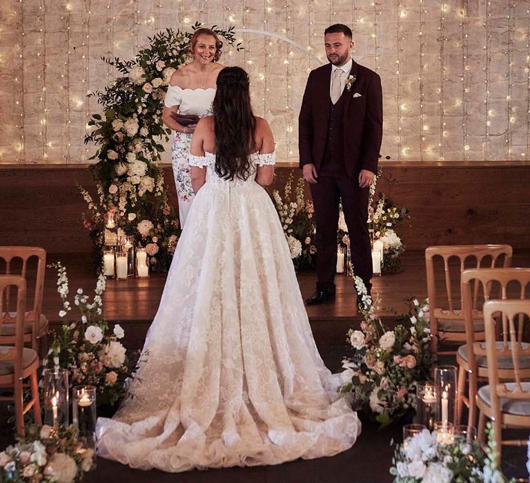 rustic barn wedding ceremony entrance with fairy light backdrop, groom in a burgundy suit and bride in an of the shoulder lace wedding dress