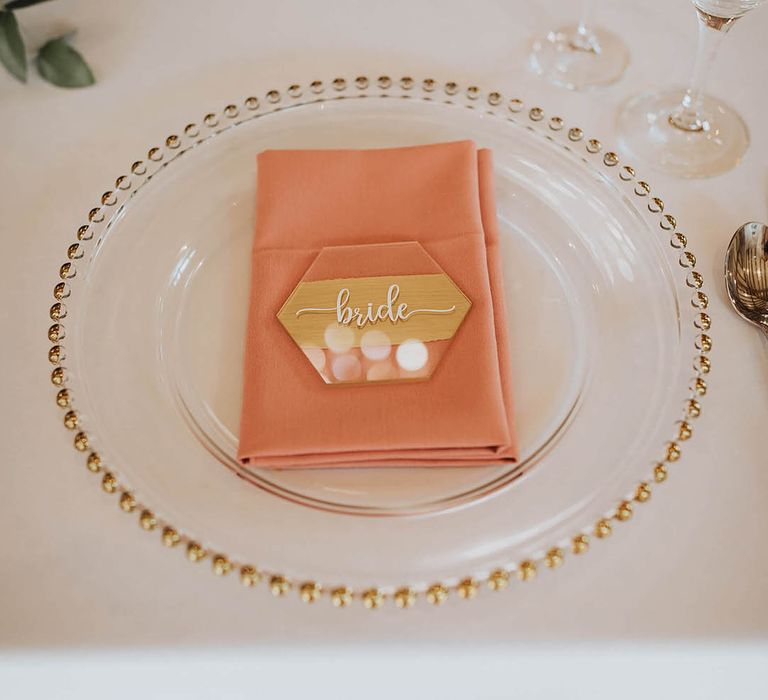 Gold edged clear charger plates with a pink napkin and hexagonal place name 