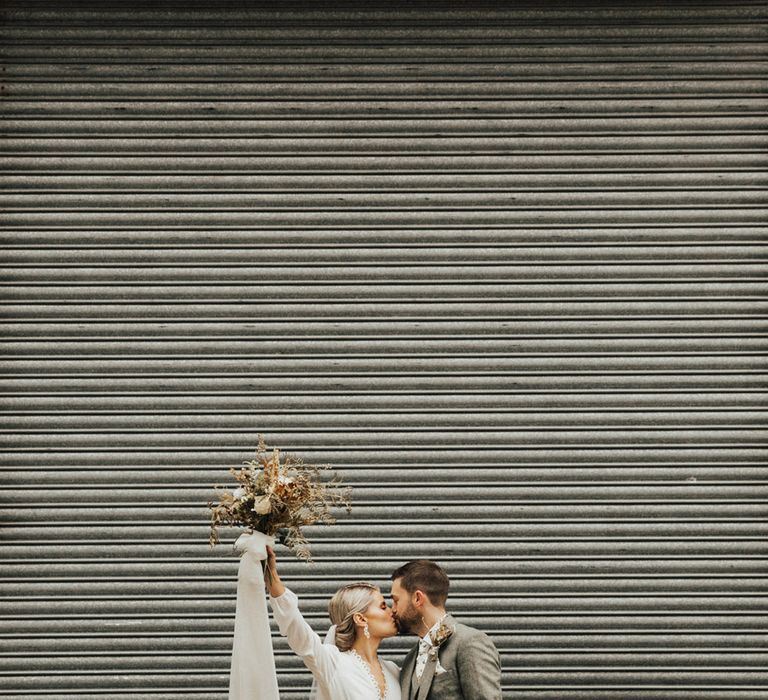 Bride holds up dried floral bouquets with white ribbon and stands with her groom in three piece grey suit