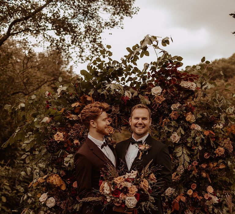 Grooms stand smiling in front of their autumnal flower arch and holding a matching bouquet 