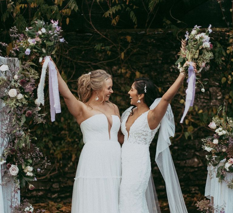 Brides in short sleeve v neck wedding dress with puff lace shoulders and sleeveless ruched wedding dress holding up mixed floral bouquets with with white carnations, purple tulips, roses and foliage and lilac and white lace ribbon