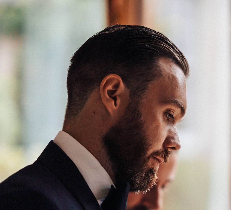 The best man with dark hair slicked back with a full beard in a tuxedo for the country house wedding 