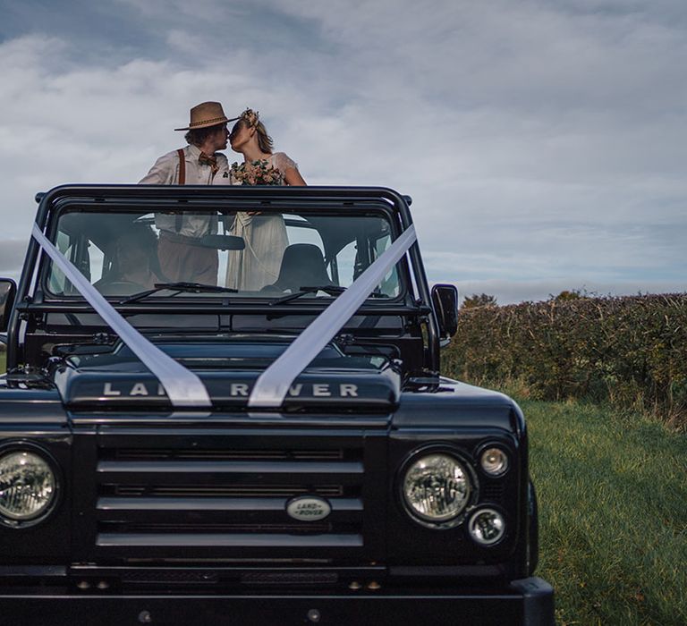 The bride and groom kiss as they stand together in the back of Land Rover decorated with white ribbon 