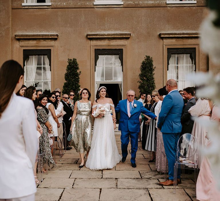 Bride in off the shoulder wedding dress is walked down the aisle by her mother and father with the bride waiting at the end of the aisle 