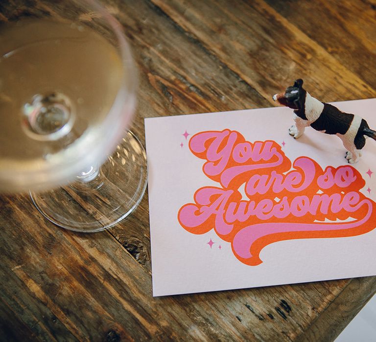 Pink, orange and red purple wedding stationery with small plastic toy dog on top next to a glass of champagne