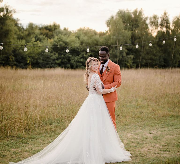 Groom in burnt orange rust coloured suit with brown tie, pocket square, brown accessories and dried flower, rose and pampas grass boutonniere hugging bride in long sleeved v neck soft ivory dress with lace floral detailing on the bodice and sheer sleeves at a harvest wedding 