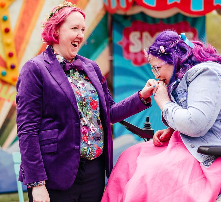 Bride in purple velvet blazer and black suit trousers with purple bowtie, pink patterned pocket square and colourful patterned shirt smiling and laughing with bride in wheelchair wearing personalised denim jacket, pink wedding dress, blue tights and gemstone bridal crown 