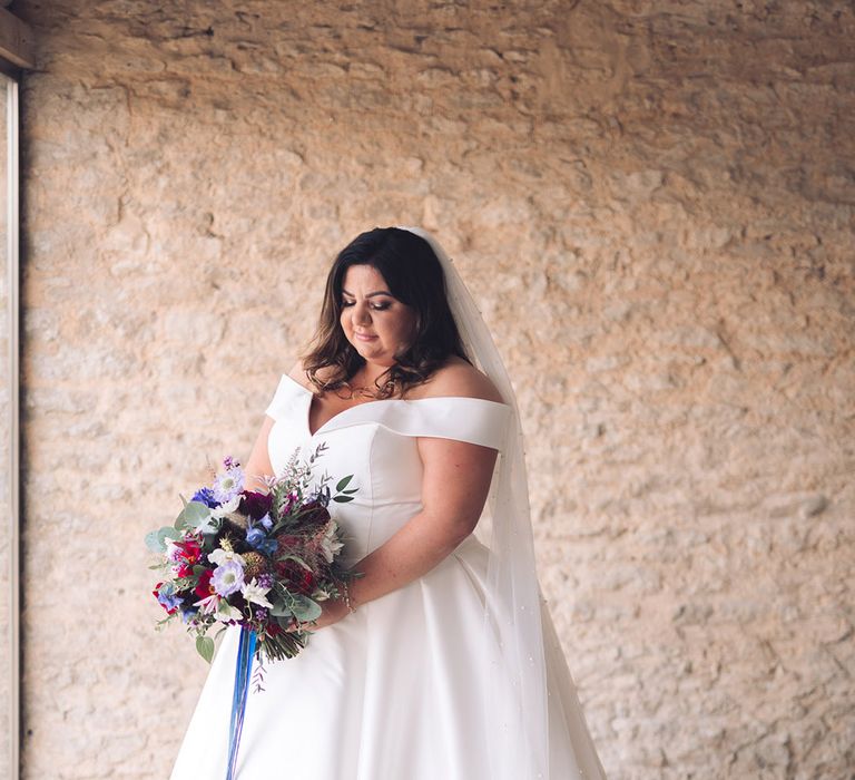 Bride with soft curled brown hair in an off the shoulder wedding dress with pearl veil holding a blue, purple, and red bouquet with blue ribbon 