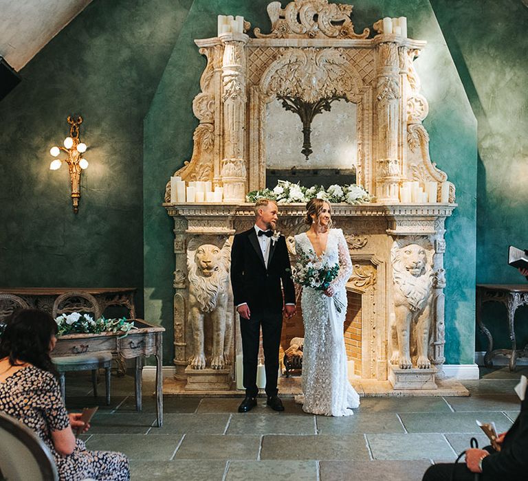 Groom in traditional black tuxedo standing with the bride in a 3D lace wedding dress with shoulder pads standing at the altar for their wedding ceremony 