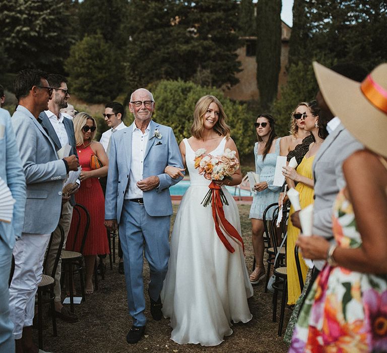 Bride in Suzanne Neville wedding dress walks down the aisle with her father who wears blue suit and white shirt 