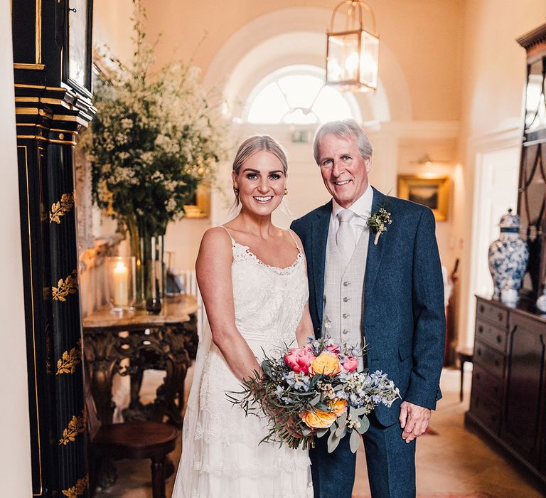 The father of the bride wears a blue and silvery grey three piece suit standing with the bride in a fitted lace ruffle wedding dress 