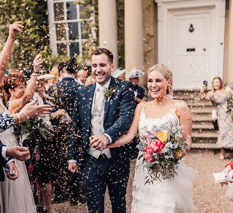 The groom wears a blue suit and grey waistcoat walking along with the bride in a fitted wedding dress for their confetti exit after the ceremony 