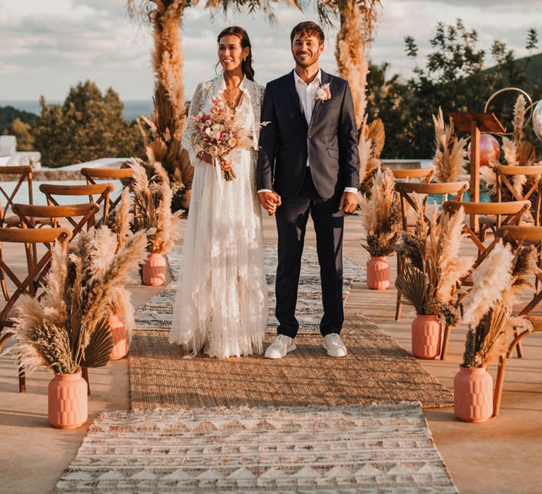 Pampas grass and dried palm leaves in orange vases line the aisle for an outdoor boho wedding 