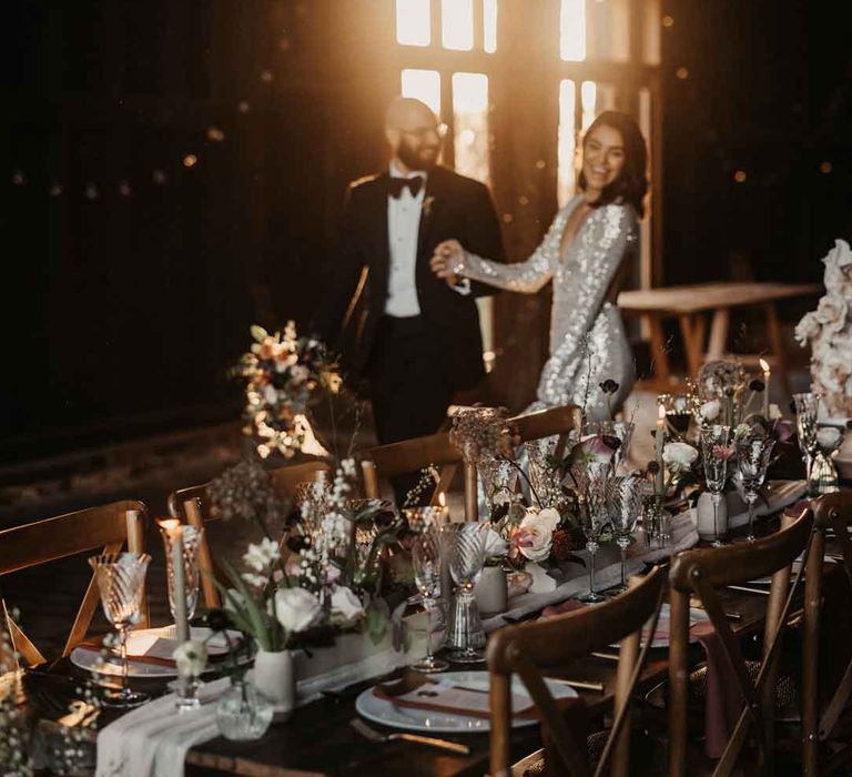 Bride in silver sparkly wedding dress and groom in classic black tuxedo with black bow tie walking by rustic luxe wedding tablescape 