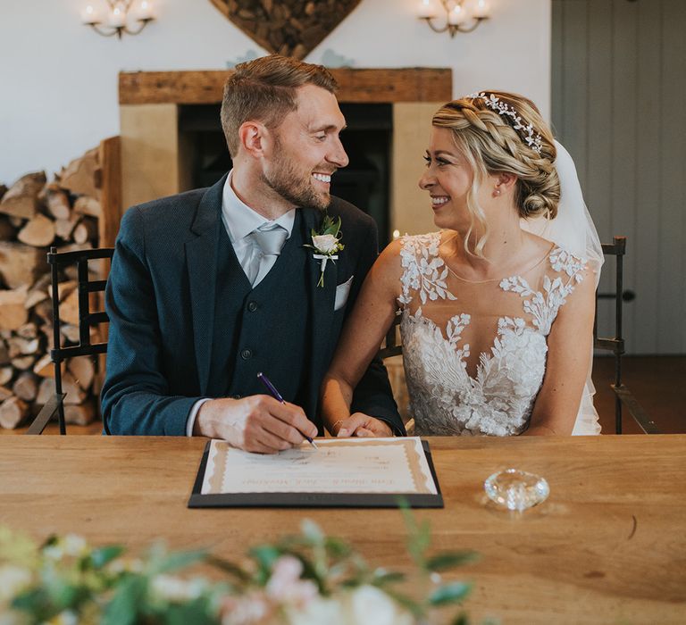 Groom in dark navy three piece suit with silver grey tie smiles at the bride as they sign the wedding register together 