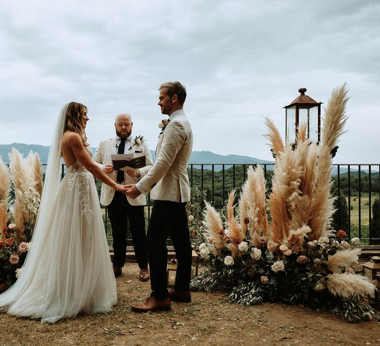 Bride in white embroidered tulle dress holds grooms hands at Italian wedding ceremony surrounded by pampas grass and white, orange and pink rose decor