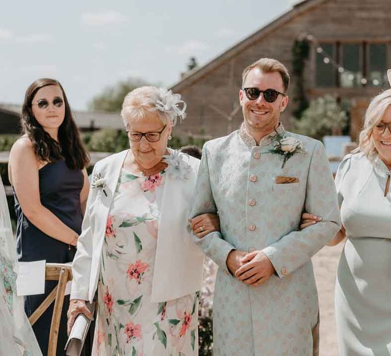 Groom in mint green Indian Sherwani with white garden rose and foliage boutonniere and grooms sunglasses walking down the aisle with family members 