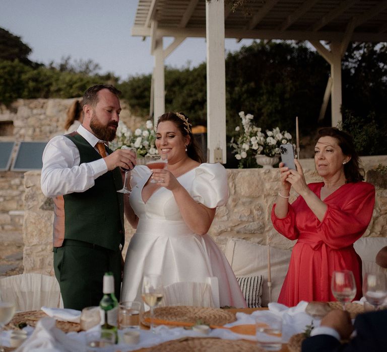 Bride in white satin wedding dress cheersing the groom in a dark green suit with a cocktails and red wine at private villa wedding