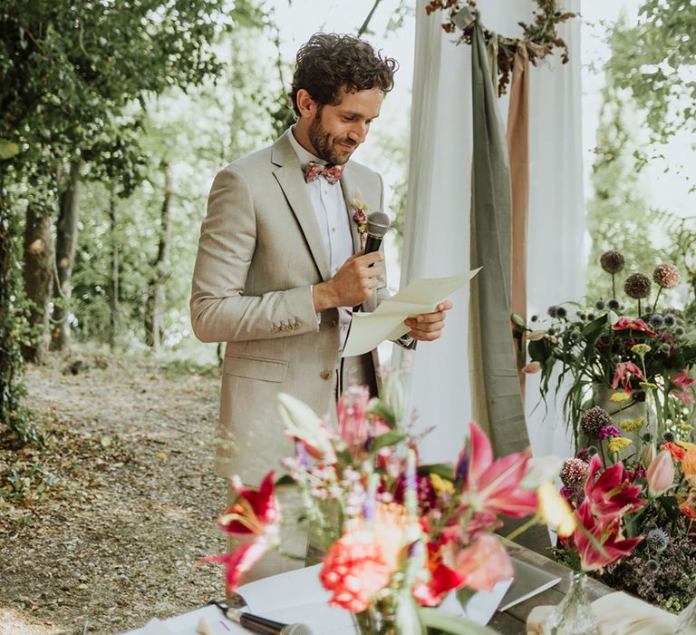 Groom in griege suit and colourful bow tie reads his written words to the bride during Italian woodland wedding ceremony