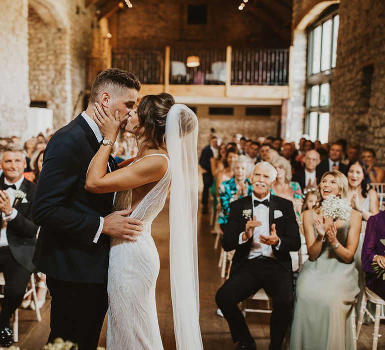 The bride in a beaded wedding dress and groom in black tie share their first kiss as a married couple 