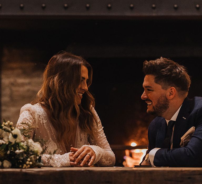 The bride in a sparkly wedding dress sits smiling at the groom in his suit as they sign the wedding register together