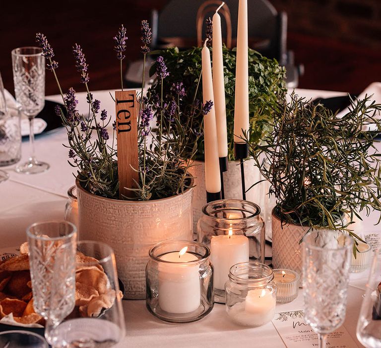 Potted plants of rosemary and lavender with a wooden table number sign along with white taper candles in black candleholder with smaller tea lights and pillar candles