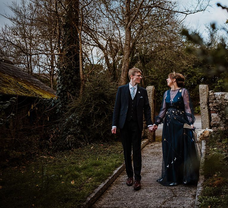 Groom in navy three piece suit walking with the bride in a navy sheer long sleeve bespoke wedding dress 