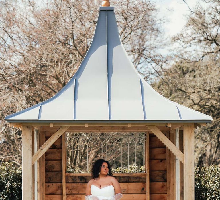 Bride in strapless wedding dress with front slit, puddle train and mesh pearl gloves standing underneath pagoda at Rackleys Barn 