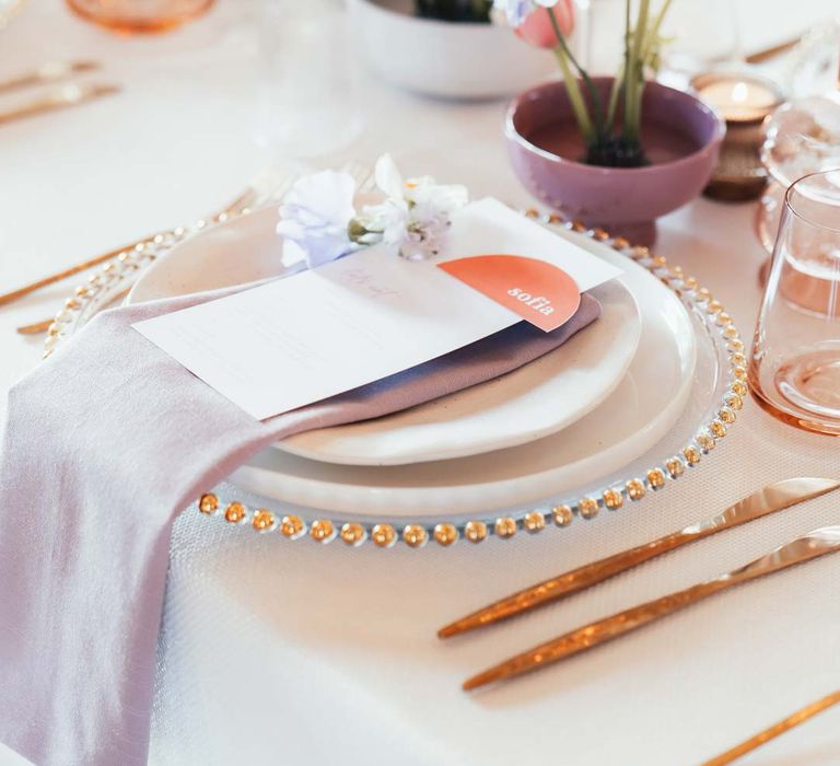 Peach and lilac wedding tablescape with spring flower centrepieces, lilac napkins, tinted glassware and gold cutlery and crockery on white tablecloth at Rackleys Barn