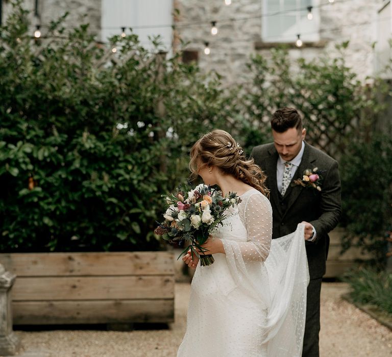 Bride with half up half Godwin hair wearing a dotted long sleeve wedding dress and veil walking with the groom in a grey suit 