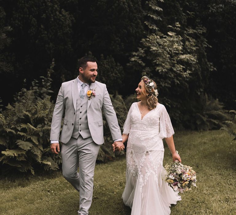 Bride in boho wedding dress walking hand in hand with the groom in a light grey wedding suit 