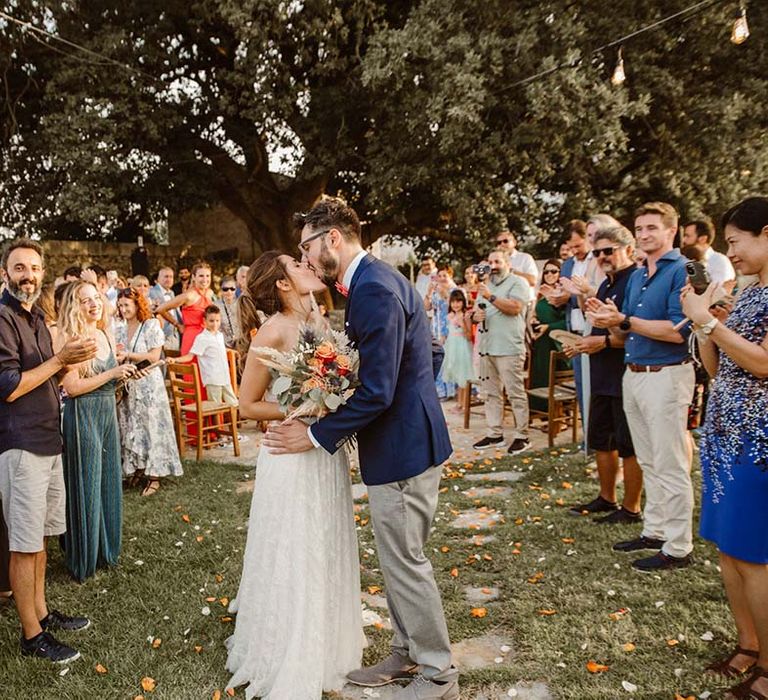 boho bride in a lace wedding dress and groom in chinos and blazer kissing at the end of their confetti exit