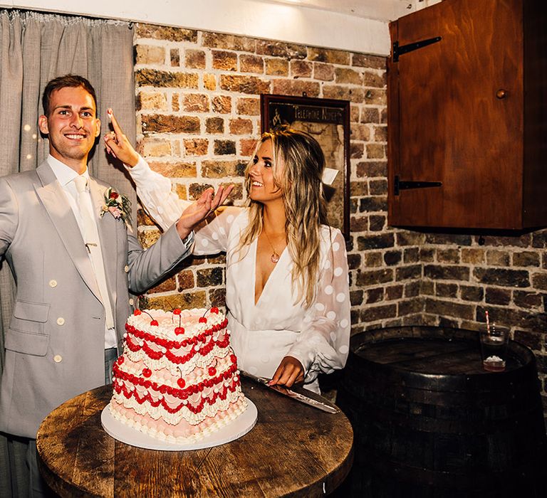 Bride in in a long sleeve bridal jumpsuit putting icing from the heart retro wedding cake on the grooms nose in a light grey wedding suit 