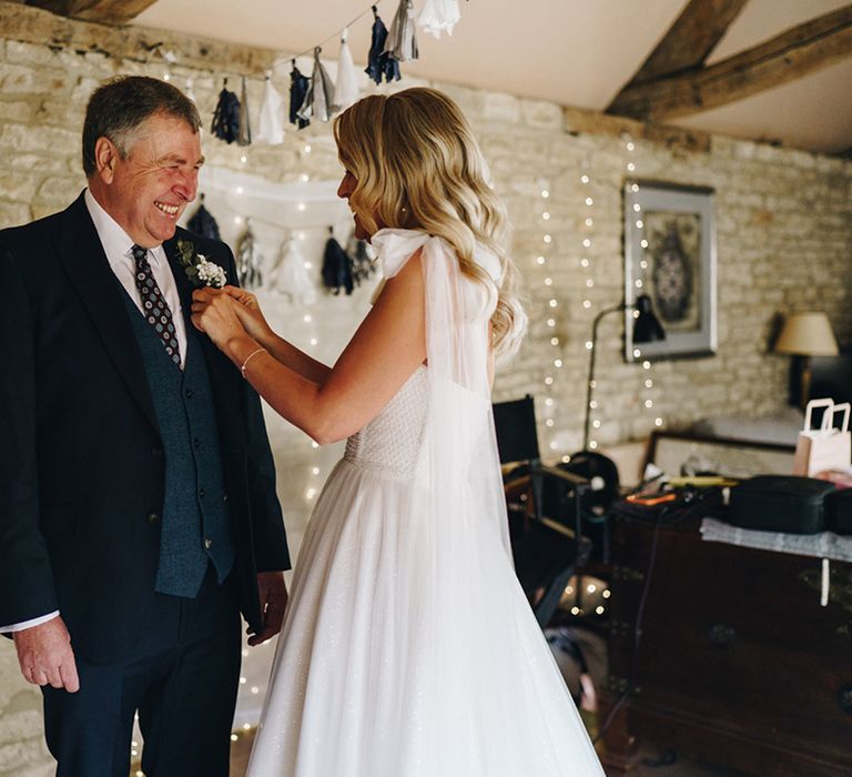 Bride with blonde wavy hair in sparkly wedding dress helps the father of the bride with his buttonhole accessory 