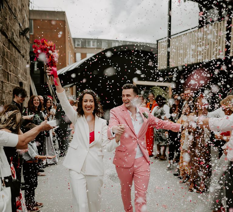 Confetti moment for the bride and groom as they exit their wedding ceremony with the bride wearing a white suit and the groom wearing a pink suit